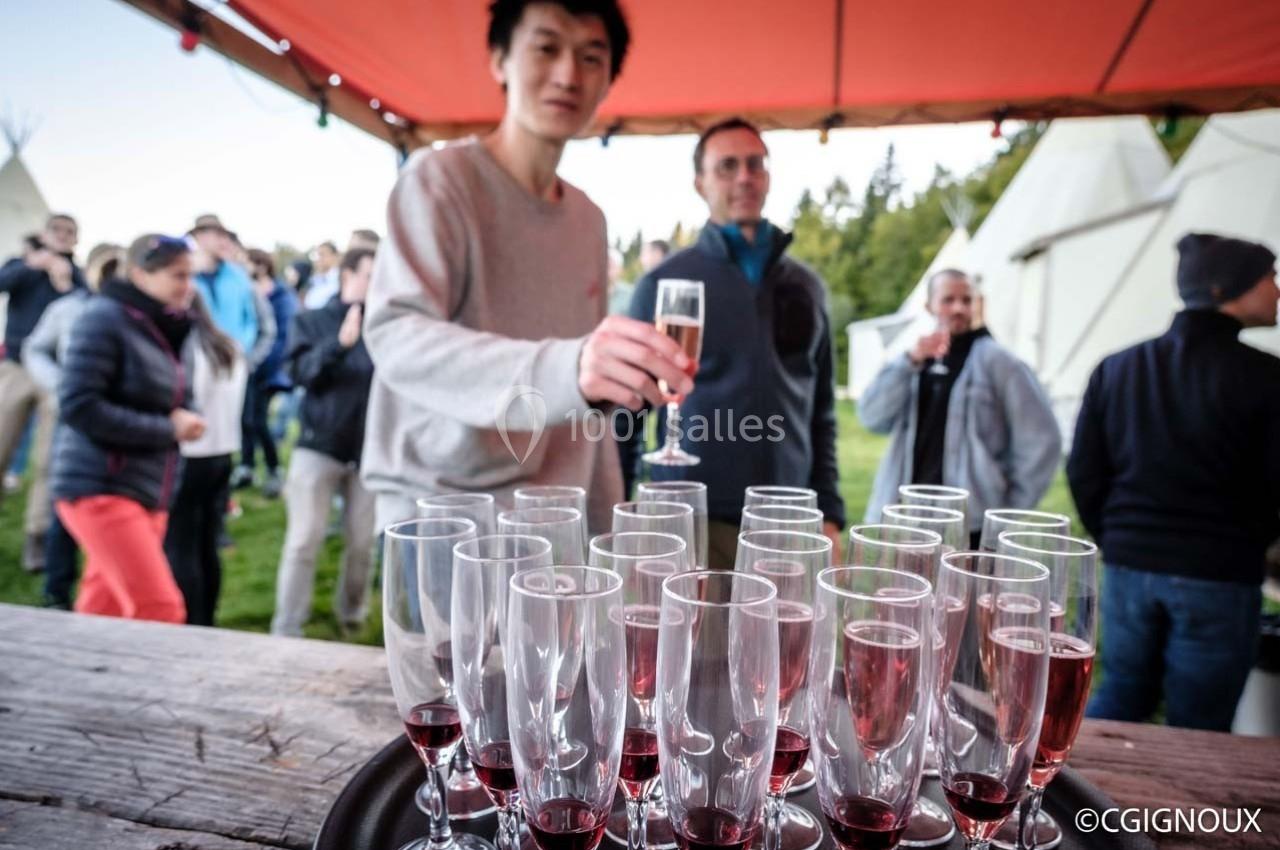 Un homme tend un verre de vin pétillant devant une table garnie de flûtes remplies, en extérieur sous une tente.