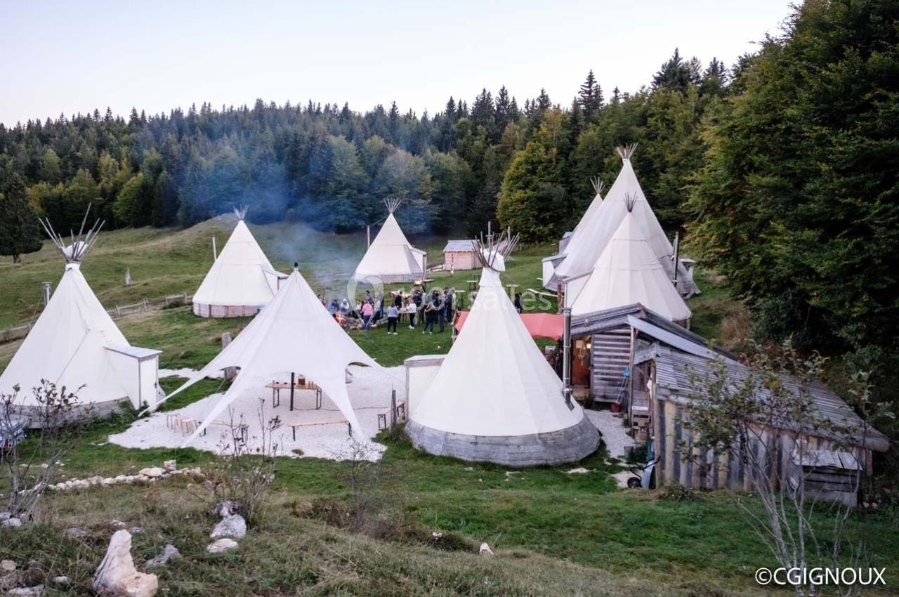 Campement de tipis et structures en bois dans une clairière entourée de forêt, avec un groupe de personnes rassemblées.