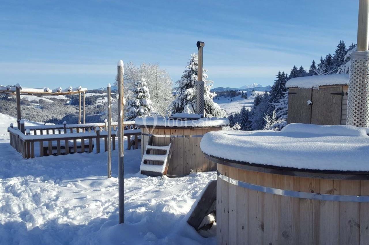 Bains nordiques en bois entourés de neige, avec vue sur des montagnes et une forêt sous un ciel bleu.