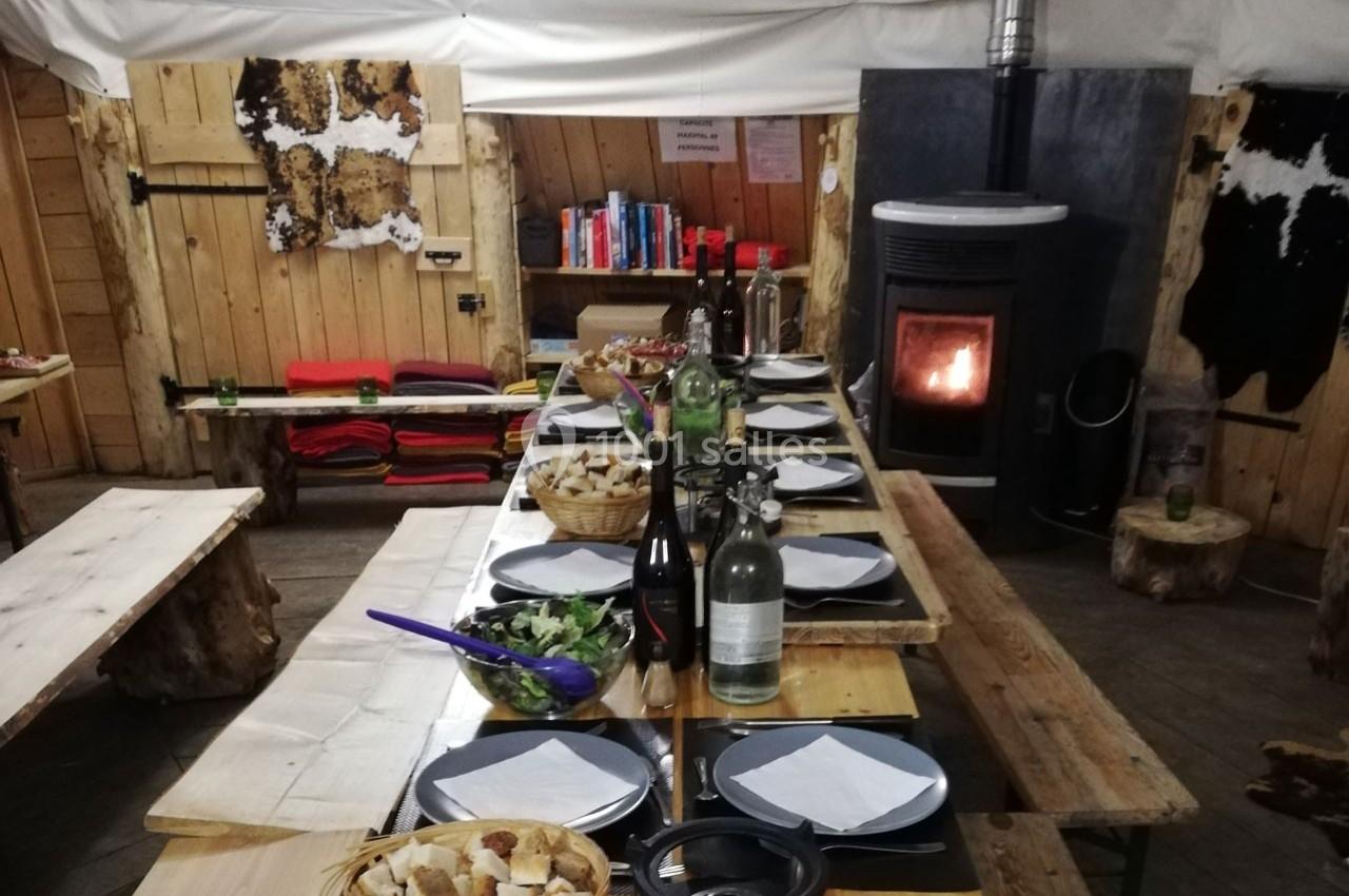 Table en bois dressée avec des assiettes, du pain et des boissons, dans une salle chaleureuse avec poêle à bois allumé.