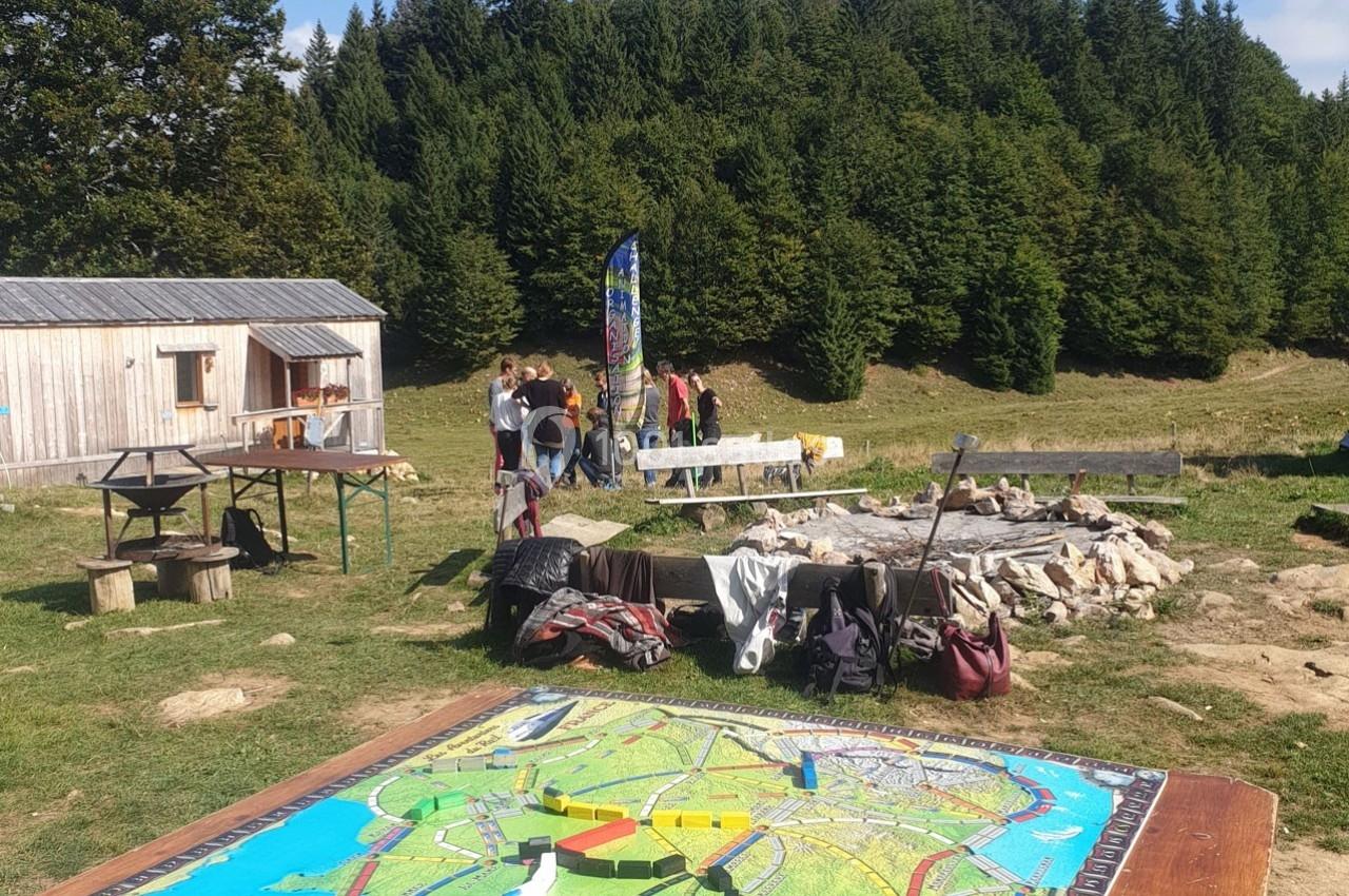Un plateau de jeu coloré au premier plan, avec un groupe de personnes près d'un feu de camp en pleine nature.