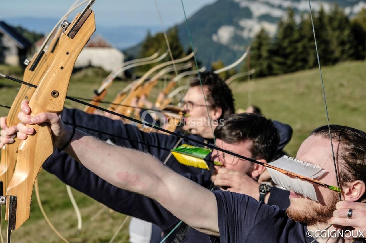 Des archers alignés tirent à l'arc en extérieur, dans un paysage de montagne verdoyant.
