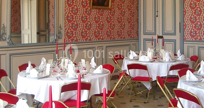 Salle élégante avec murs ornés de papier peint rouge, tables dressées avec nappes blanches et chaises rouges pliantes.
