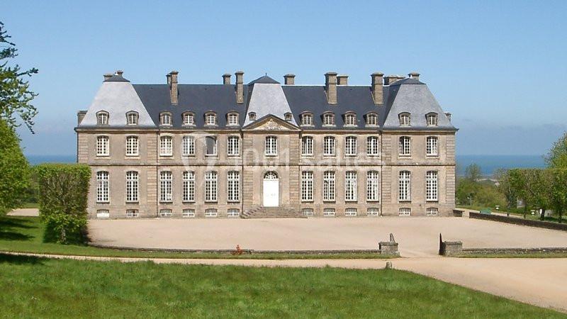 Façade d'un château en pierre avec toit en ardoise, entouré de verdure et vue sur la mer à l'arrière-plan.
