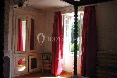 Chambre lumineuse avec lit à baldaquin, armoire à miroir, chaise en bois et rideaux rouges encadrant une fenêtre ouverte.