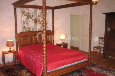 Chambre lumineuse avec lit à baldaquin, armoire à miroir, chaise en bois et rideaux rouges encadrant une fenêtre ouverte.