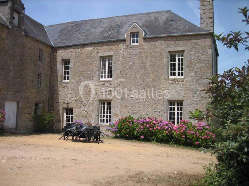Façade en pierre d'une maison ancienne avec des fenêtres blanches, hortensias en fleurs et une table de jardin noire.