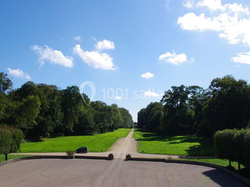 Allée bordée d'arbres avec une voiture garée sur le côté, sous un ciel bleu parsemé de nuages.