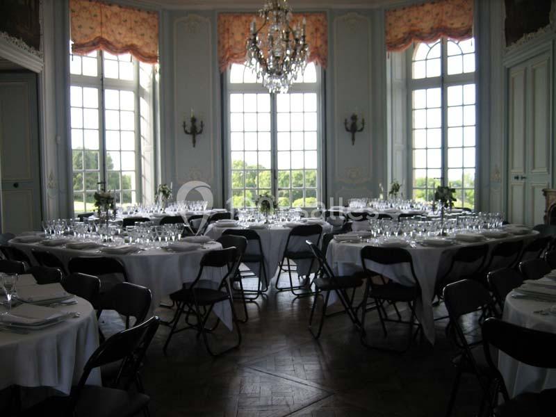 Salle de réception élégante avec tables rondes dressées, nappes blanches, chaises noires et grandes fenêtres donnant sur un…
