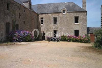 Vue d'un village avec des maisons entourées de verdure, un chemin en premier plan et la mer à l'horizon sous un ciel bleu.
