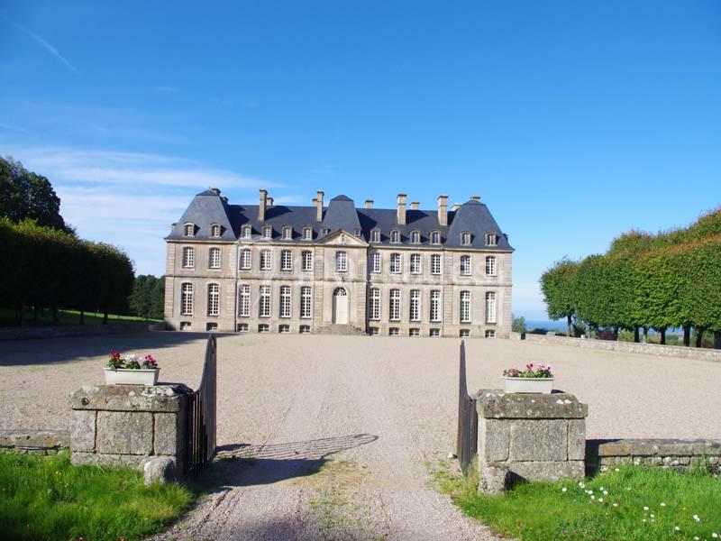 Façade d'un château en pierre avec toit en ardoise, entouré d'arbres et d'une allée gravillonnée.