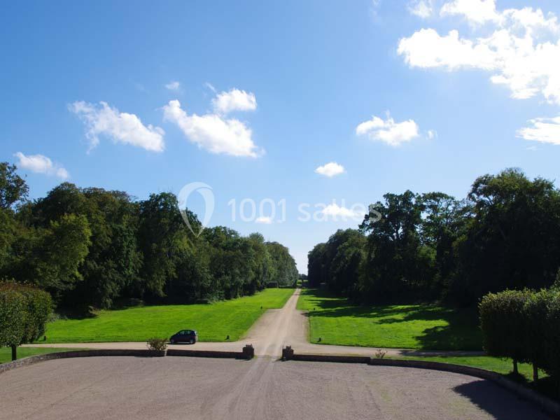 Allée bordée d'arbres et de pelouses verdoyantes sous un ciel bleu avec quelques nuages, une voiture garée à gauche.