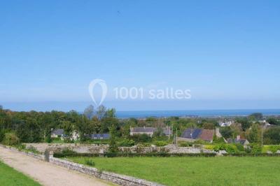 Vue d'un village avec des maisons entourées de verdure, un chemin en premier plan et la mer à l'horizon sous un ciel bleu.