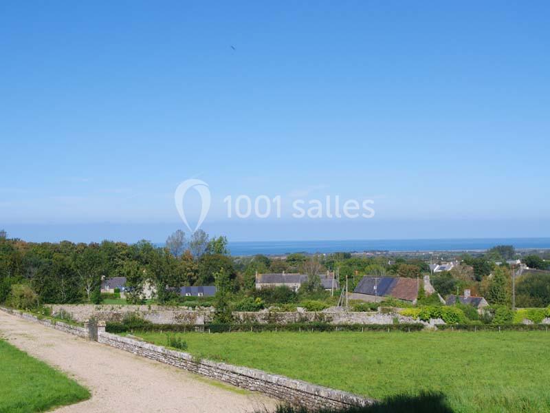 Vue d'un village avec des maisons entourées de verdure, un chemin en premier plan et la mer à l'horizon sous un ciel bleu.