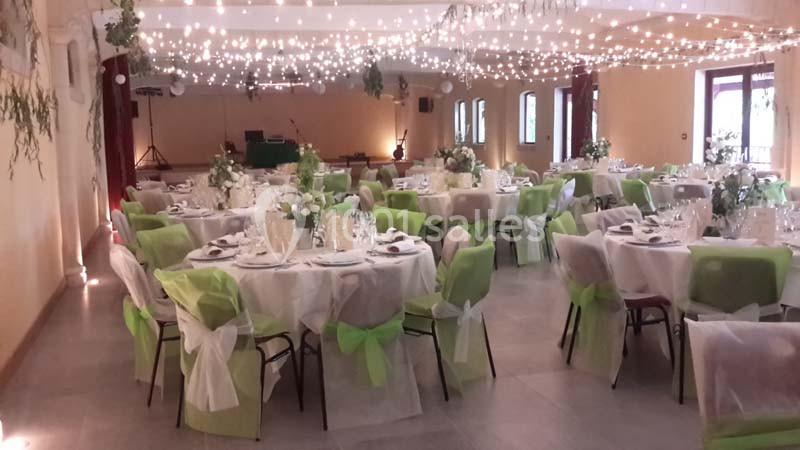 Salle de réception décorée avec des tables rondes, nappes blanches, chaises ornées de rubans verts et guirlandes lumineuses.