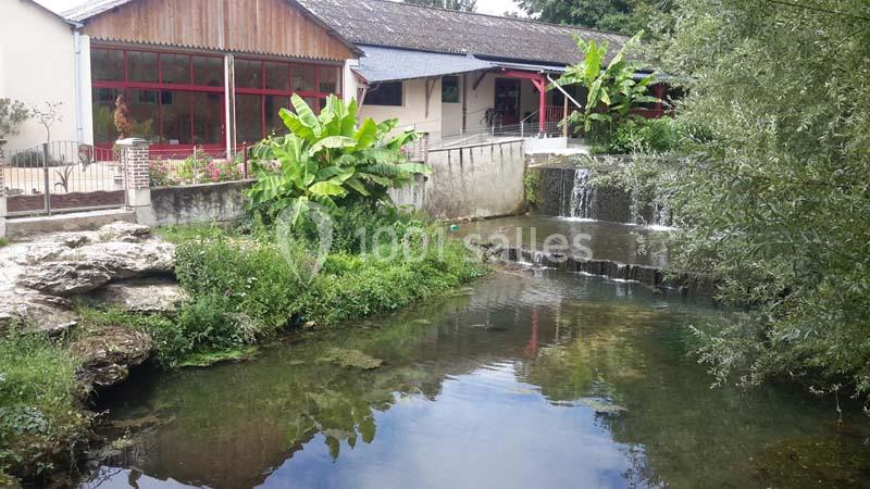 Vue d'un bâtiment avec terrasse près d'une rivière, entouré de végétation et d'une petite cascade.