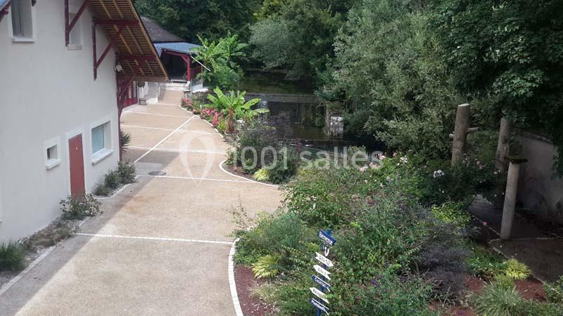 Chemin bordé de végétation menant à un bâtiment clair, avec un petit cours d'eau et des arbres en arrière-plan.