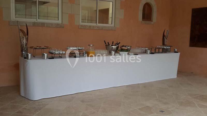 Table de buffet dressée avec boissons, verrines et couverts, dans une salle aux murs couleur pêche et fenêtres encadrées de…