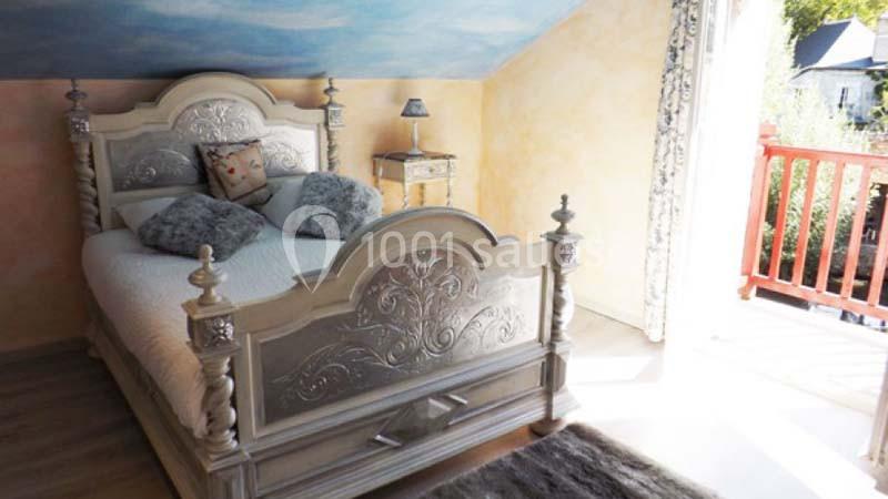 Chambre lumineuse avec lit double sculpté, murs jaunes, plafond peint en ciel et balcon ouvert sur l'extérieur.