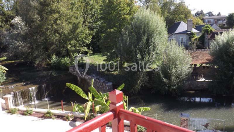 Vue depuis une terrasse rouge sur un petit cours d'eau bordé d'arbres, avec des maisons en arrière-plan.