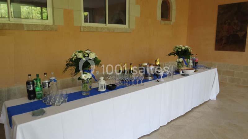 Table dressée avec nappes blanches, verres, bouteilles, seau à champagne et décorations florales dans une salle lumineuse.
