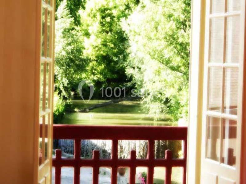 Vue depuis une fenêtre ouverte sur un balcon rouge, donnant sur un jardin verdoyant et un plan d'eau en arrière-plan.