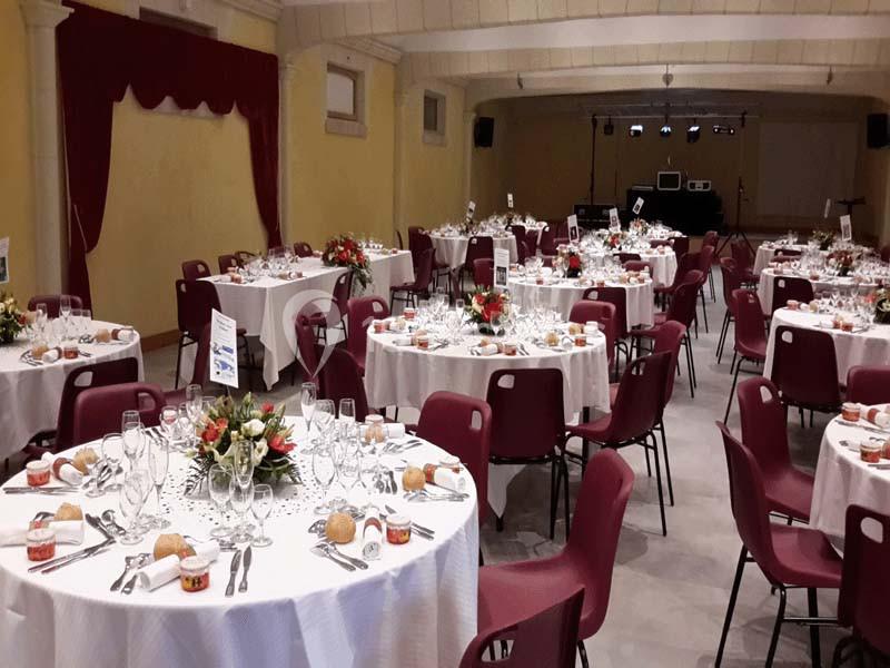 Salle de réception avec des tables rondes dressées pour un repas, décorées de fleurs et entourées de chaises rouges.