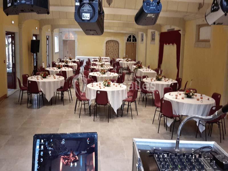 Salle de réception avec tables rondes dressées, chaises rouges et équipements audiovisuels en premier plan.