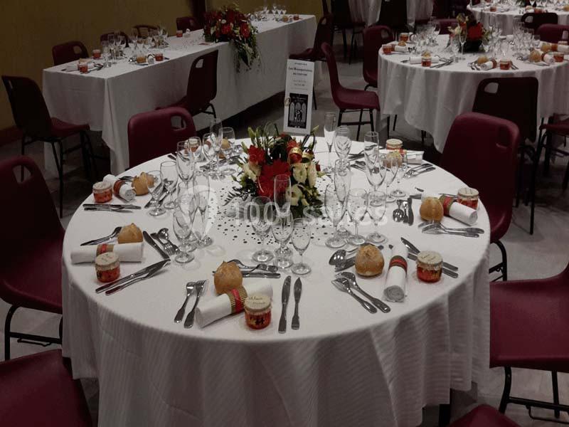 Table ronde dressée pour un repas, avec nappes blanches, couverts, verres, décorations florales et petits pains.