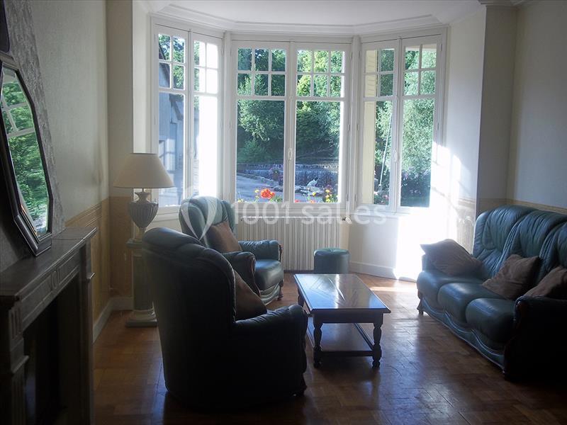 Salon lumineux avec fauteuils en cuir vert, table basse en bois et grande baie vitrée donnant sur un jardin verdoyant.