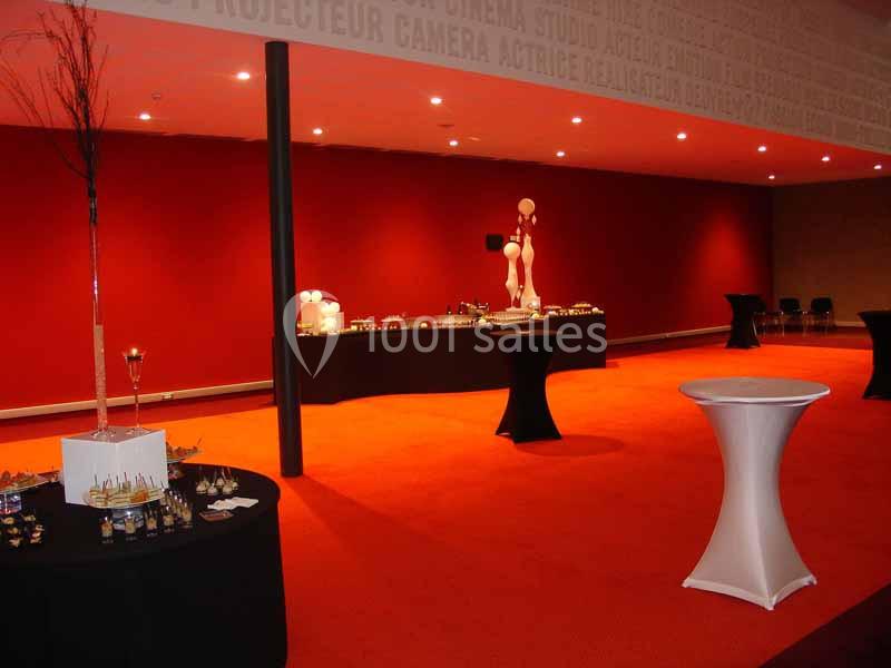 Salle de réception avec moquette rouge, tables hautes et buffet dressé en arrière-plan.