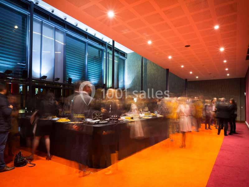 Personnes rassemblées autour d'une table de buffet dans une salle éclairée avec un sol orange et des murs sombres.