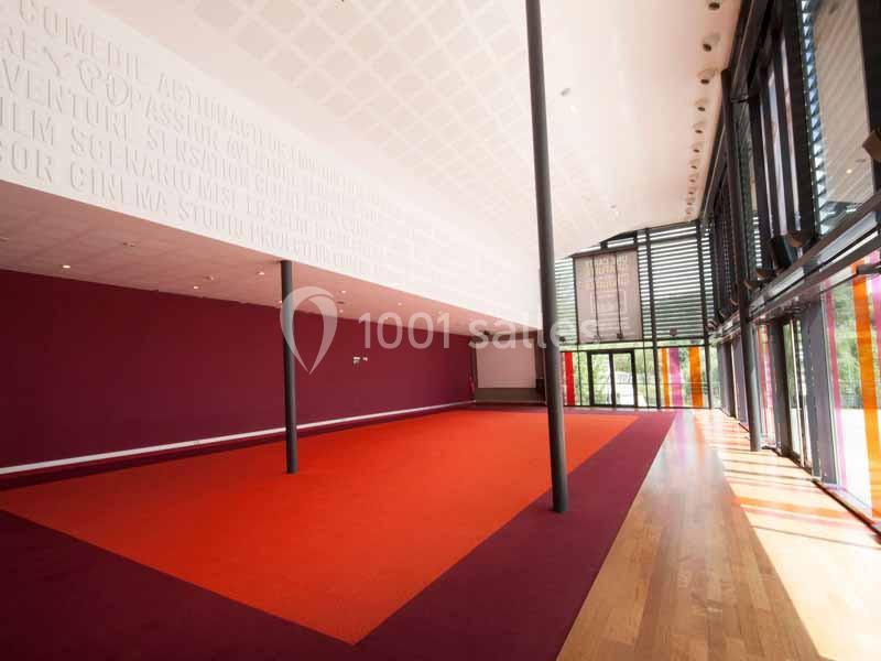 Salle lumineuse avec murs rouges, sol en moquette orange et parquet, grandes baies vitrées laissant entrer la lumière…