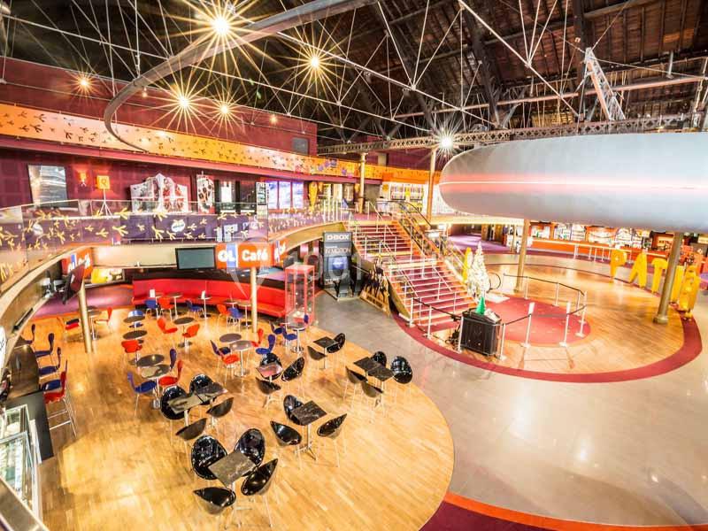 Vue intérieure d'un espace de loisirs avec sièges, tables, escalier central et décorations colorées sous un plafond haut.