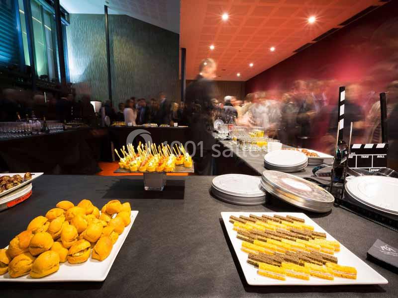 Buffet avec amuse-bouches variés sur une table, des invités flous en arrière-plan dans une salle éclairée.