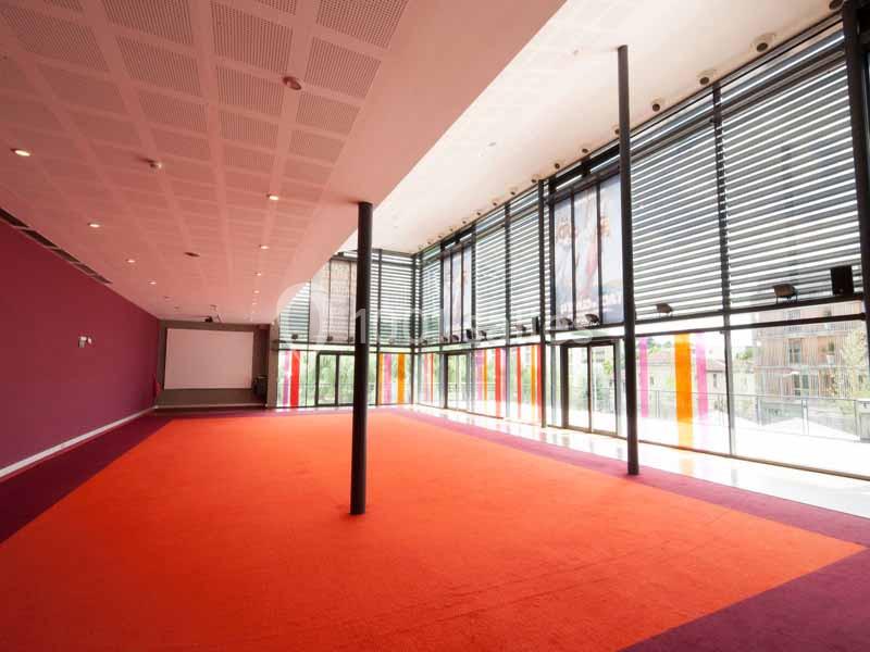 Salle lumineuse avec de grandes baies vitrées, stores horizontaux et moquette rouge-orange.