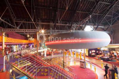 Vue intérieure d'un espace de loisirs avec sièges, tables, escalier central et décorations colorées sous un plafond haut.