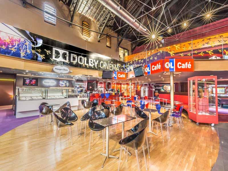 Intérieur d'un cinéma avec espace café, tables, chaises modernes et enseignes lumineuses sous un plafond industriel.