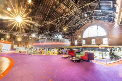 Vue intérieure d'un espace de loisirs avec sièges, tables, escalier central et décorations colorées sous un plafond haut.