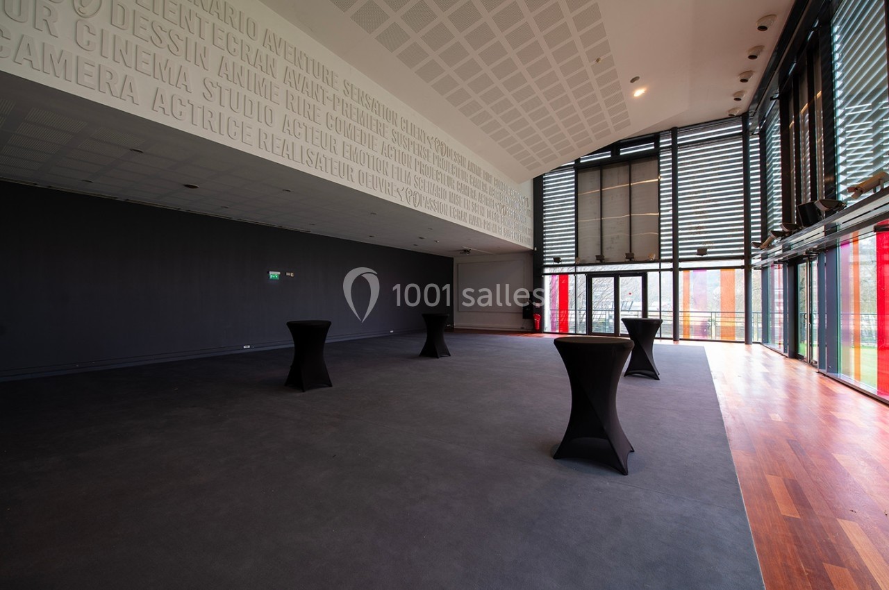 Salle vide avec sol en moquette, tables hautes noires, baies vitrées et murs décorés de mots en relief.