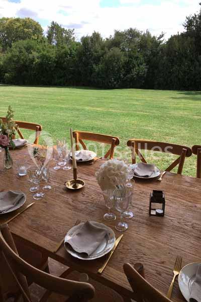 Table en bois dressée avec vaisselle élégante, bougies et fleurs, donnant sur une pelouse et des arbres en arrière-plan.