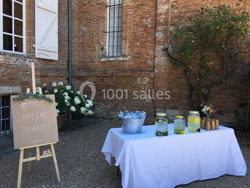Table dressée en extérieur avec boissons fraîches, pots en verre et panneau en bois près d'un mur en briques.