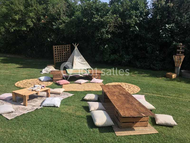 Aménagement extérieur avec tente tipi, tables en bois, coussins et tapis sur une pelouse entourée de végétation.