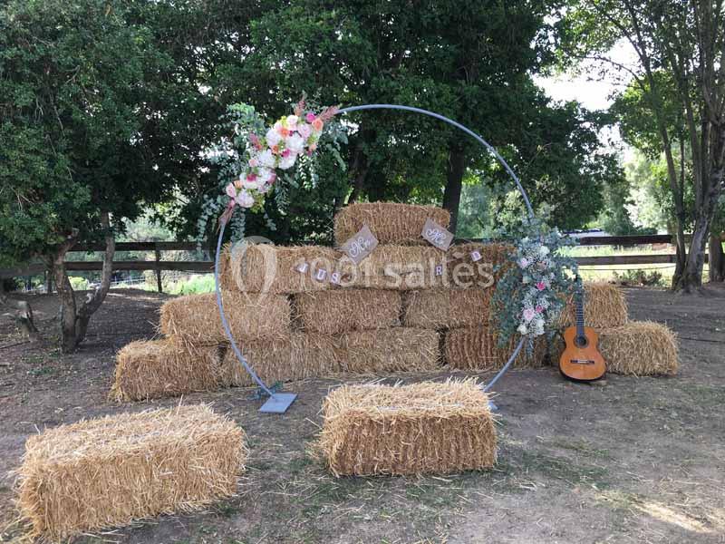 Bottes de paille empilées décorées de fleurs, guirlandes et une guitare dans un cadre extérieur arboré.