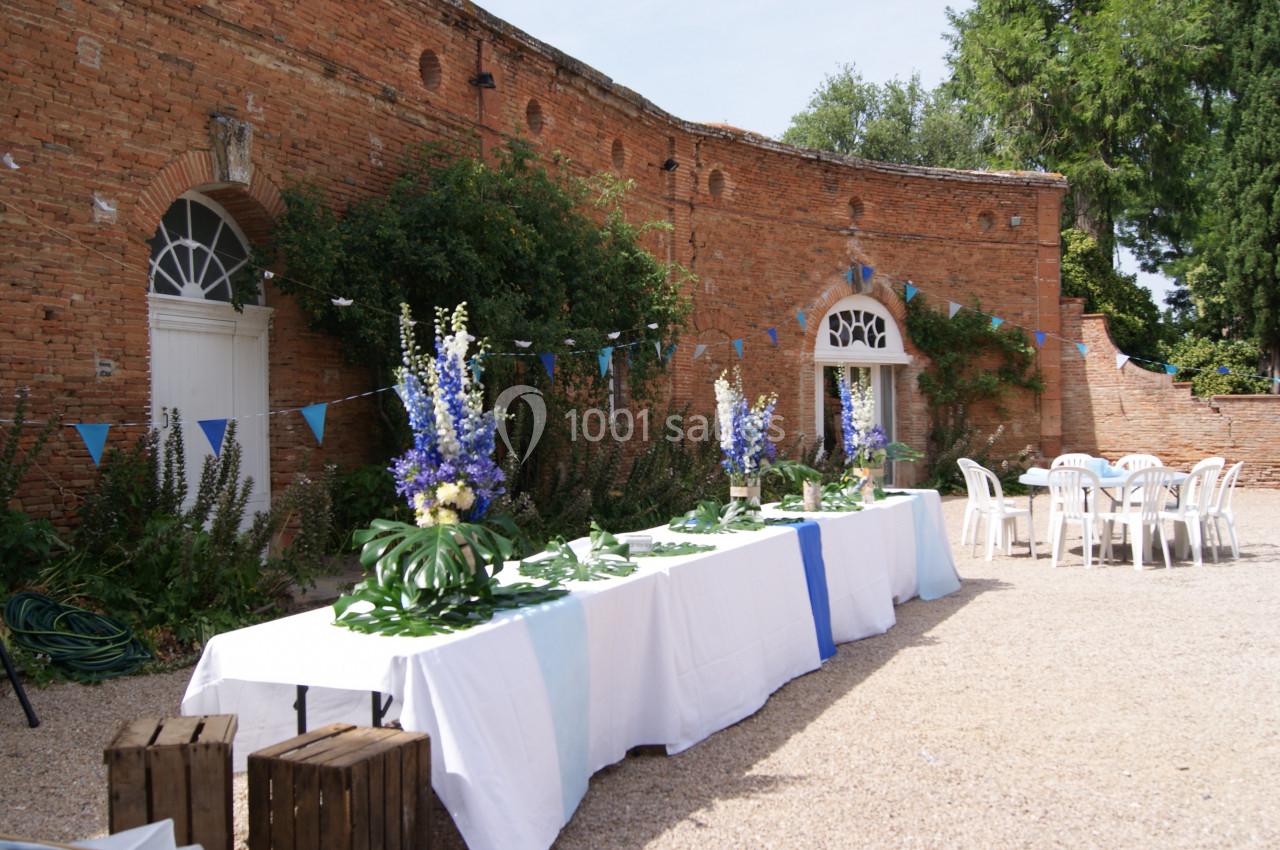 Tables décorées de nappes blanches et fleurs bleues dans une cour en briques, avec chaises blanches et guirlandes.
