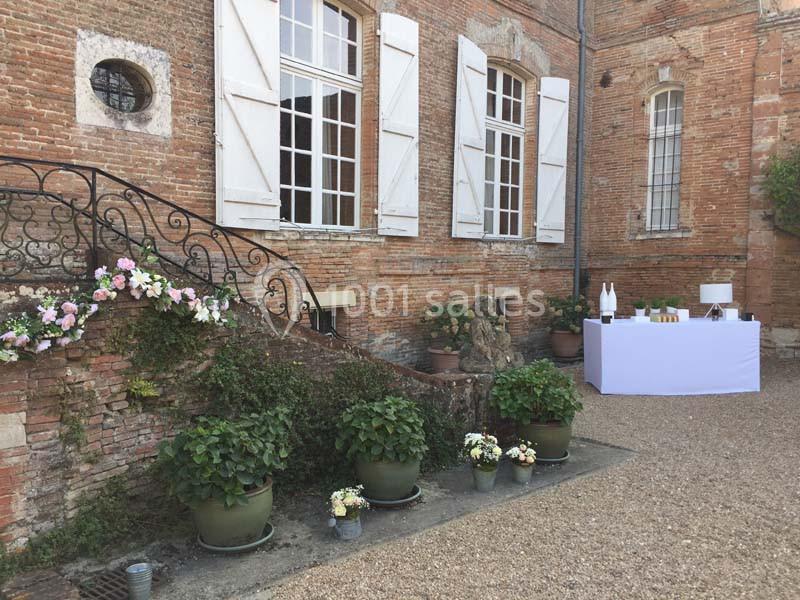 Façade en briques avec fenêtres à volets blancs, escalier fleuri et table dressée dans une cour gravillonnée.
