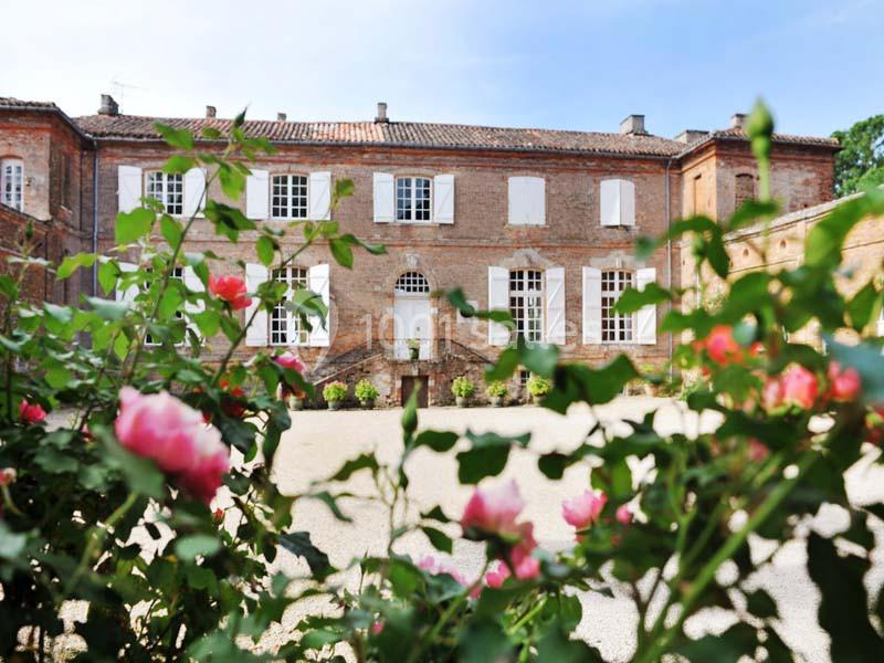Façade d'un bâtiment en briques rouges avec des fenêtres blanches, vue à travers des rosiers en fleurs au premier plan.