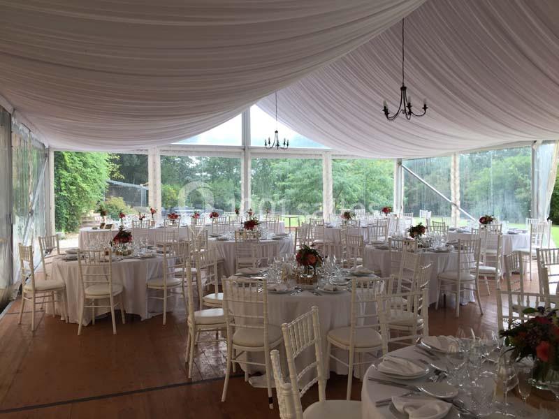 Salle de réception sous une tente avec tables rondes dressées, chaises blanches et vue sur un jardin verdoyant.