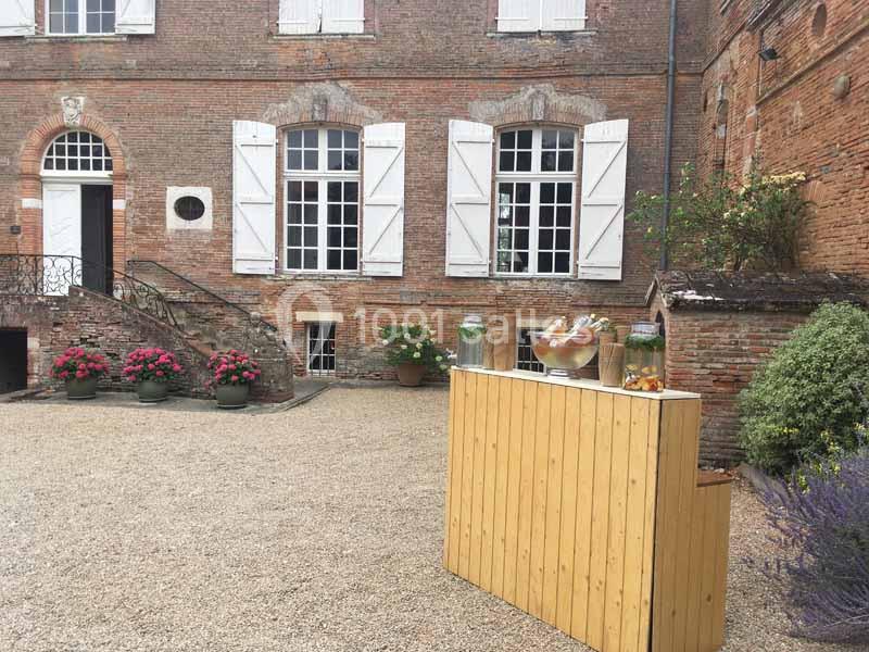 Façade en briques d'un bâtiment ancien avec fenêtres à volets blancs, terrasse gravillonnée et bar en bois au premier plan.