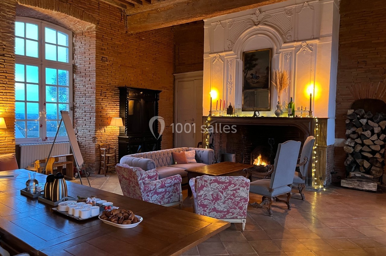 Salon chaleureux avec cheminée allumée, mobilier ancien, table dressée et murs en briques apparentes.